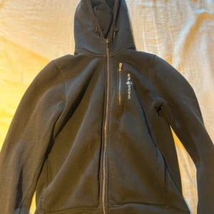 Svart hoodie från Sail Racing - Säljer en svart hoodie från Sail Racing i bra skick. Den har en dragkedja framtill och en praktisk ficka med dragkedja på bröstet. Perfekt för höst och vår, med en skön huva för extra värme. Passar både till vardags och lite sportigare aktiviteter.