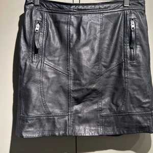 Svart skinnkjol från AllSaints - Säljer en snygg svart skinnkjol från AllSaints i storlek 40. Kjolen har en cool bikerstil med dragkedja bak och fickor fram. Perfekt för en edgy look till både vardag och fest. Tillverkad i 100% läder med viskosfoder för extra komfort.