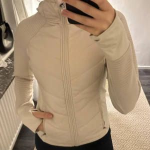 Beige jacka med huva - Säljer en snygg beige jacka med huva och dragkedja. Den har långa ärmar med ribbade detaljer och är perfekt för höst och vår. Jackan har en figurnära passform och är i mycket bra skick. Perfekt för vardagsbruk eller en promenad!