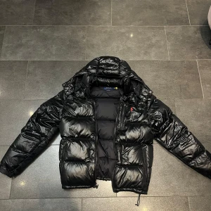 Svart pufferjacka från Polo Ralph Lauren Glossy - Säljer en snygg svart pufferjacka från Polo Ralph Lauren i storlek M. Jackan är perfekt för kalla dagar med sitt tjocka, vadderade material och har en dragkedja framtill. Den är i nyskick, inga defekter eller ett enda tecken på använding, passar perfekt för både höst och vinter. Köpt för 6095kr från Zalando, kvitto finns!