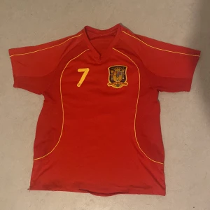 Spanien fotbollströja  - Säljer en röd fotbollströja med gula detaljer och nummer 7 på framsidan. Tröjan har korta ärmar och ett emblem på bröstet. På baksidan står det 'David Villa' med samma nummer. Perfekt för fotbollsfans som vill visa sitt stöd! 🏆