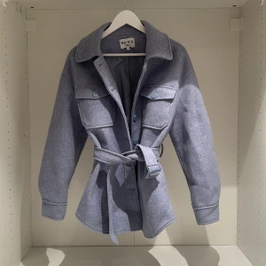 Ljusblå/lila jacka från NA-KD - Säljer en superfin ljusblå/lila jacka från NA-KD i storlek 34. Jackan har långa ärmar och ett bälte i midjan för en snygg passform. Den har också stora fickor framtill och knappar för stängning. Perfekt för vår och höst!