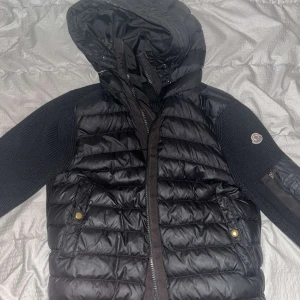 Svart cardigan från Moncler - Säljer en snygg svart cardigan från Moncler. Jackan har en quiltad design med stickade ärmar och en praktisk huva. Den är perfekt för höst och vinter och har en dragkedja framtill samt fickor med knappar. Moncler-loggan syns på ärmen. Perfekt för dig som vill hålla stilen i kylan!