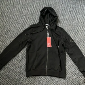 Svart huvtröja från C.P. Company - Säljer en svart huvtröja från C.P. Company i storlek M, men den sitter som storlek S. Tröjan har en dragkedja framtill och en cool detalj på ärmen. Perfekt för höst och vår, med en stilren look som passar till det mesta. Nästan helt ny med tag kvar! Kan gå ner i pris vid snabb affär!