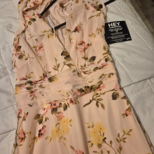 Blommig långklänning från Hey - Säljer en superfin blommig långklänning från Hey i ljusrosa med vackra blommor i rosa, gul och grön. Klänningen har en omlott-design och är ärmlös, perfekt för sommarens fester eller midsommarfirandet. Den har en elegant passform som framhäver figuren. 🌸