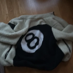 Beige och svart fleecejacka från Stussy (reverseable - Säljer en supermjuk och varm fleecejacka från Stussy i beige och svart. Den har en cool 8-ball design på ryggen och en praktisk dragkedjeficka fram. Perfekt för höst och vinter när du vill hålla stilen och värmen. 🖤🤍