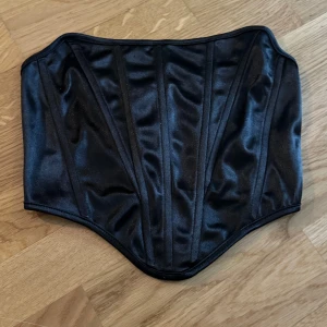 Svart korsett - Snygg svart korsett i glansigt material. Perfekt för en festlig look eller som en statement piece. Den har en klassisk design med en tight passform som framhäver midjan. Ett måste för garderoben!