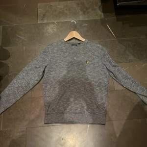 Grå tröja från Lyle & Scott - Säljer en stilren grå tröja från Lyle & Scott i mycket bra skick. Den har en klassisk design med rund hals och ett litet gult logomärke på bröstet. Perfekt för både vardag och lite finare tillfällen. Passar bra till jeans eller chinos!