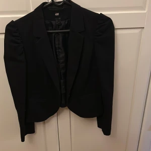 Svart kavaj från H&M - Snygg svart kavaj från H&M i storlek 42. Mycket liten i storlek. Mer som 38/40. Den har en klassisk design med puffiga axlar och långa ärmar. Perfekt för både jobb och festliga tillfällen. Kavajen är i mycket bra skick och ger en stilren look.