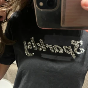 Svart Monki t-shirt med glittrig text - Säljer en svart t-shirt från monki med texten 'Sparkly' i glittriga stenar på framsidan. Perfekt för en casual look med lite extra bling! T-shirten har en normal passform och korta ärmar. Nästan aldrig använd, jättebra skick!