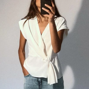 Vit omlottblus från Zara - Säljer en stilren vit omlottblus från Zara. Den har en elegant knytning i sidan och korta ärmar. Perfekt för både vardag och fest! Blusen är i mycket bra skick och passar till alla tillfällen.