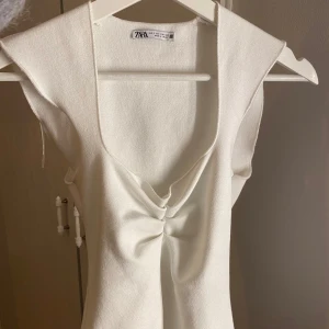 Vit topp från Zara - Säljer en stilren vit ärmlös topp från Zara. Den har en djup v-ringning och en snygg knutdetalj framtill. Toppen är i ett mjukt och bekvämt material.