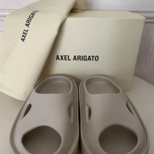 Beige sandaler från Axel Arigato - Säljer ett par stilrena beige sandaler från Axel Arigato. De är i nytt skick och oanvända, kommer med originalpåse och låda. Perfekta för sommaren med en modern design och bekväm passform. Passar 41-43 i storlek.