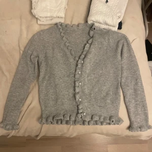 Grå stickad kofta med volangdetaljer - Superfin grå stickad kofta med volangdetaljer längs knäppningen och nederkanten. Perfekt för kyliga dagar och ger en söt touch till vilken outfit som helst. Den har långa ärmar och är i mycket bra skick och endast använd ett fåtal gånger.