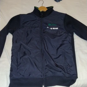 Svart jacka från Hugo Boss - Säljer en snygg svart jacka från Hugo Boss i mycket bra skick. Jackan har en dragkedja framtill och är långärmad. Perfekt för höst och vår med sin stilrena design och bekväma passform. Märkesloggan är broderad på bröstet. Passar både till vardags och lite finare tillfällen.