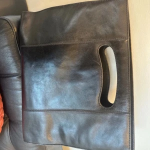 Svart skinnväska från Tiger - Snygg svart skinnväska från Tiger med praktiskt handtag och en stilren design. Väskan har en ficka med spänne på framsidan och flera fack inuti, inklusive ett med dragkedja. Perfekt för både vardag och jobb!