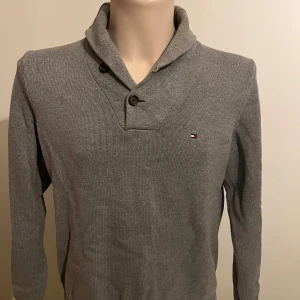 Grå tröja från Tommy Hilfiger - Säljer en stilren grå tröja från Tommy Hilfiger med en unik krage och två knappar. Tröjan har långa ärmar och en liten logotyp på bröstet. Perfekt för en avslappnad men snygg look.