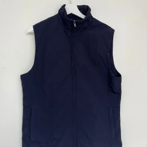 Mörkblå väst från Riley - Säljer en stilren mörkblå väst med dragkedja framtill. Västen har en hög krage och två fickor på sidorna. Perfekt för lager-på-lager under kyligare dagar.