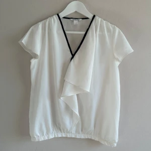 Kortärmad v-ringad blus med volang   - Stilren kortärmad blus med volang framtill från SuiteBlanco. Vit med svart kant vid v-ringning. Gott begagnat skick. Några små märken i tyget framtill men jag tycker inte det stör så mkt. Blusen är perfekt under en kavaj och då syns det inte. Försökte ta bild på det. Rökfritt hem. Rensar garderoben så kika gärna på övriga annonser! 