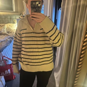 Beige och svart randig stickad tröja - Säljer en mysig beige stickad tröja med svarta ränder och dragkedja vid halsen. Tröjan har långa ärmar och ribbade muddar vid ärmslut och nederkant. Perfekt för kyliga dagar!