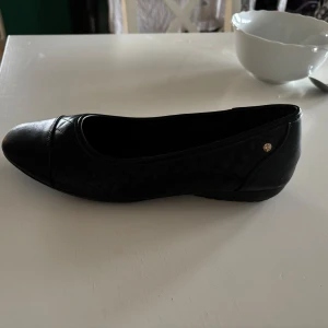 Svarta ballerinaskor - Snygga svarta ballerinaskor med en elegant design. Skorna har en rund tå och en liten metallknapp på sidan som detalj. Perfekta för en stilren look.