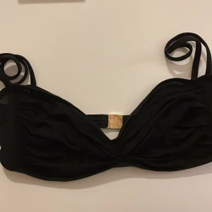 Svart bikiniöverdel från Louis Vuitton - Elegant svart bikiniöverdel från Louis Vuitton med axelband och en stilren gulddetalj. Perfekt för en lyxig dag vid poolen eller stranden. 