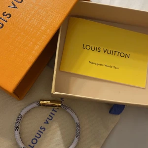 Armband från Louis Vuitton - Elegant armband från Louis Vuitton med vit och svart design. Armbandet har guldfärgade detaljer och kommer i originalförpackning med en Louis Vuitton-påse. Perfekt för att ge en lyxig touch till din stil.
