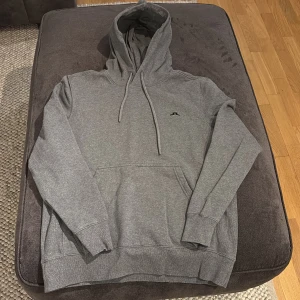 Grå hoodie från J.Lindeberg - Säljer en stilren grå hoodie från J.Lindeberg. Den har en klassisk design med en känguruficka och justerbar huva med dragsko. Perfekt för en avslappnad look.