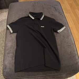 Svart pikétröja från Boss - Snygg svart pikétröja från Boss med grå detaljer på krage och ärmslut. Tröjan har en klassisk pikédesign med knappar framtill och Boss-logga på bröstet. Perfekt för en stilren look.