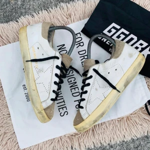 Vita sneakers med svarta snören - Snygga vita sneakers från Golden Goose med svarta snören och beige detaljer. Skorna har en klassisk design med en stjärna på sidan och en något sliten look för en trendig stil.