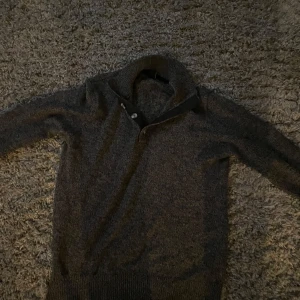 Mörkgrå stickad tröja med krage - Säljer en stilren mörkgrå stickad tröja med krage och knappdetalj. Perfekt för kyligare dagar med sin mjuka och bekväma passform. Tröjan har långa ärmar och ribbstickade muddar vid ärmslut och nederkant.