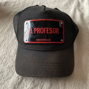 Svart keps från John Hatter & Co - Snygg svart keps med texten 'El Profesor' i rött på en metallplatta framtill. Kepsen har ett nätmaterial baktill för bättre ventilation och justerbar passform. Perfekt för en cool och avslappnad stil. Solblekt men annars fin