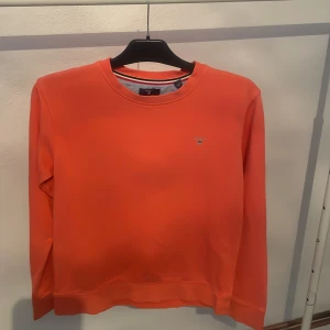 Orange tröja från GANT - Säljer en snygg orange tröja från GANT med broderad logga på bröstet. Tröjan har en klassisk rund hals och långa ärmar, perfekt för en stilren look. Passar både till vardags och mer avslappnade tillfällen.