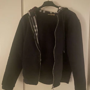 Svart hoodie med dragkedja från burberry - Säljer en svart hoodie från Burberry med dragkedja och klassiskt rutigt mönster i huvan. Perfekt för en casual look. Den har långa ärmar och en bekväm passform. Använd ganska mycket men är ändå i typ 7/10 skick. Priset är inte hugget i sten