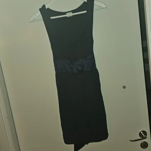 Svart ärmlös klänning med knytband - Elegant svart ärmlös klänning med ett dekorativt knytband i midjan som knyts ihop baktill. Perfekt för en stilren look.