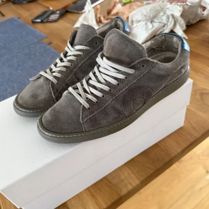Oliver Cabell - Snygga grå sneakers i mocka med vit snörning och en cool blå detalj vid hälen. Skorna har en stilren design med en diskret logga på sidan. Perfekta för en avslappnad stil. Dessa Oliver Cabell kostar 3000 plus tull och mitt pris är 1200 så det är en bra deal. Inga skador på skorna förutom att man ser att de är använda vid hälen. 