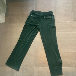 Gröna velourbyxor från Juicy Couture - Snygga gröna velourbyxor från Juicy Couture med snörning i midjan och fickor fram. Perfekta för en avslappnad stil med en touch av lyx. Matcha med den tillhörande hoodien för en komplett look. Ej några defekter, original pris ligger på 1800.