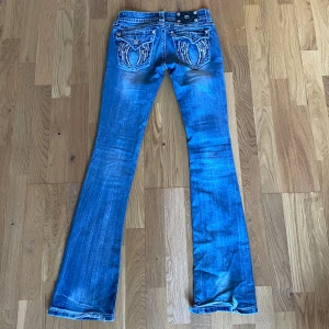 Blå jeans från Miss Me - Snygga blå jeans från Miss Me med bootcut-stil. De har dekorativa broderier och nitar på bakfickorna dock är en av knapparna på bakfickan borta. Storlek 25, skriv om du har någon fråga ☺️Midjemått är ca 35 cm rakt över och innerbenslängd är ca 83 cm