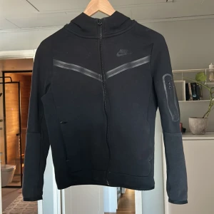  Nike tech fleece - Säljer en svart jacka från Nike med dragkedja och långärmad design. Den är lite sliten men inte mycke. Den har även gått sönder lite vid höger ficka men går att laga lätt med att sy.