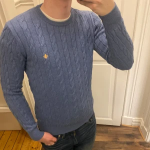 Blå kabelstickad tröja från Morris - Säljer en snygg blå kabelstickad tröja från Morris. Tröjan har en liten broderad detalj på bröstet och är perfekt för kyligare dagar. Den har långa ärmar och en klassisk rund halsringning. Passar utmärkt till jeans eller chinos för en stilren look. Modellen är ca 182, tveka inte att kontakta vid eventuella frågor 😀