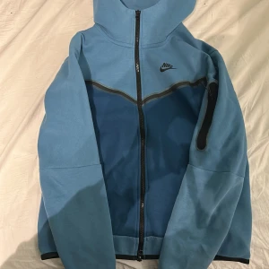 Blå hoodie nike tech (hyfsad sällsynt) - Säljer en blå hoodie från Nike med dragkedja och huva. Jackan har en ficka med dragkedja på ärmen och svarta detaljer. Perfekt för en sportig look.