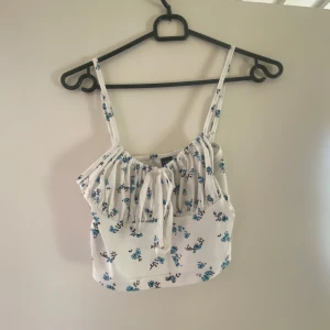 Vit blommig linnetopp - Söt vit linnetopp med blå blommor och justerbara axelband. Toppen har en knytning framtill och är perfekt för varma dagar. Den har en lätt ribbad struktur som ger en fin detalj.
