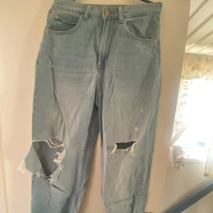 90-tals baggy jeans i ljusblå denim - Säljer ett par ljusblå 90-tals inspirerade baggy jeans med hög midja. Jeansen har slitna detaljer på knäna och en klassisk femficksdesign. Perfekta för en avslappnad stil med en vintage touch.