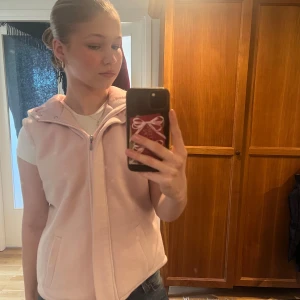 Ljusrosa fleeceväst från Next - Säljer en mysig ljusrosa fleeceväst från Next med dragkedja och huva. Perfekt för kyliga dagar när du vill hålla dig varm och bekväm. Västen har praktiska fickor framtill.