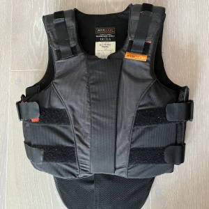 Svart säkerhetsväst från Airowear - Säljer en svart säkerhetsväst från Airowear, perfekt för ridning. Västen är i storlek T1 Slim Regular och har en dragkedja framtill. Den är designad för att ge maximalt skydd och komfort med justerbara remmar för en perfekt passform. Västen är i nyskick och uppfyller säkerhetsstandarden EN13158:2009 Level 3.