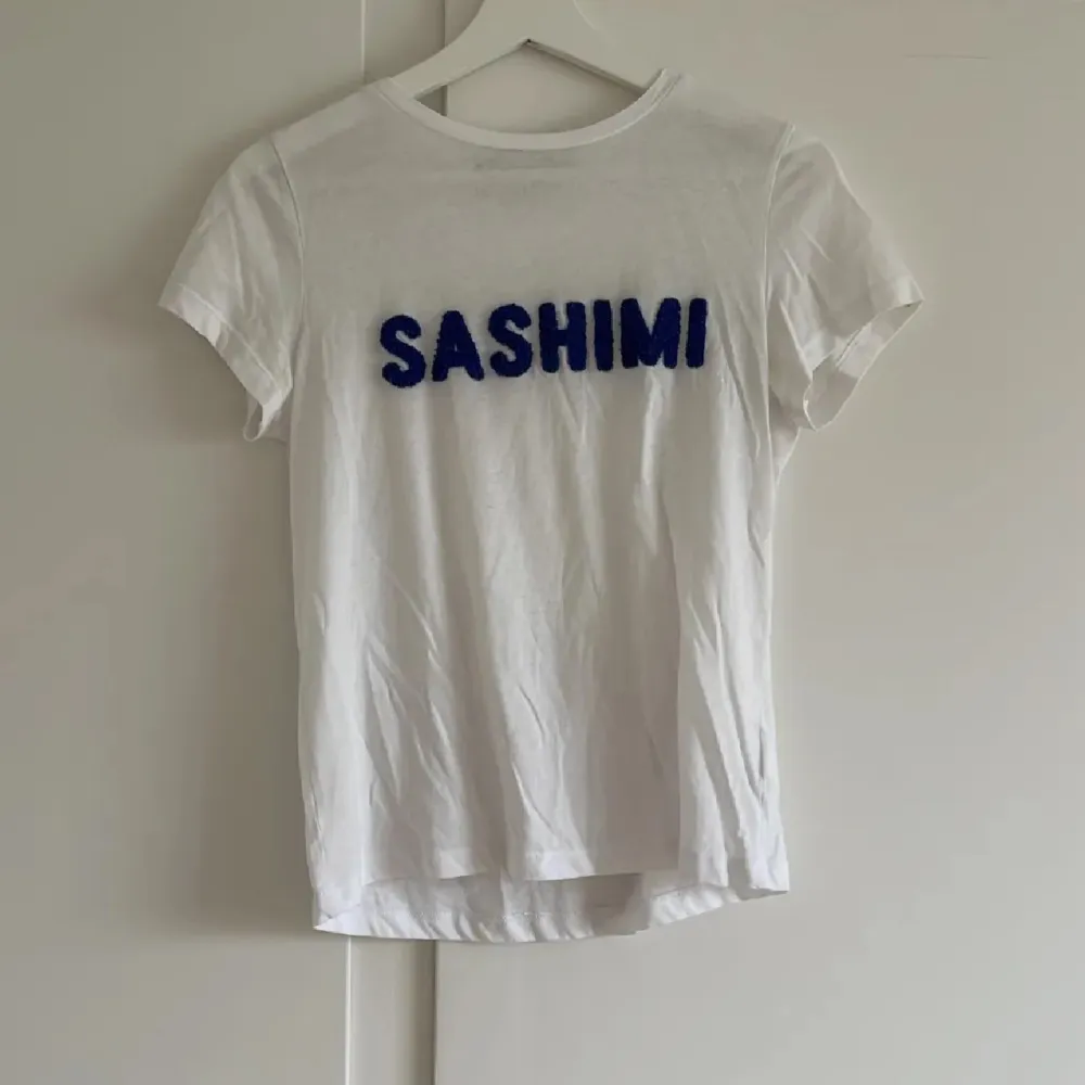 Säljer en vit t-shirt med texten 'SASHIMI' i blått på framsidan. T-shirten är kortärmad och har en normal passform. Perfekt för en avslappnad stil!. T-shirts.