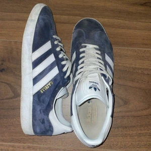 Adidas Gazelle i blå mocka - Säljer ett par klassiska Adidas Gazelle sneakers i blå mocka med vita detaljer. Skorna har den ikoniska tre-randiga designen på sidorna och en vit sula. De är i bra skick med lite slitage på sulan. Perfekta för vardag. Kommer med marinblåa snören