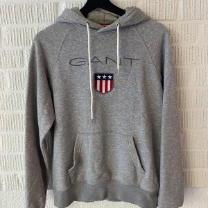 Grå hoodie från GANT - Säljer en snygg och bekväm grå hoodie från GANT i storlek S. Den har en klassisk design med GANT-loggan och ett coolt emblem på framsidan. Hoodien är perfekt för både höst och vinter, med långa ärmar och en mysig huva. Den är i bra skick😊