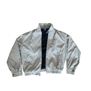 Beige jacka från Polo Ralph Lauren - Säljer en snygg beige jacka från Polo Ralph Lauren. Den har en klassisk krage och dragkedja framtill. Insidan är fodrad med ett stilrent rutigt mönster i mörkblått och grönt. Jackan är i polyester med bomullsfoder, perfekt för vår och höst. Den har också det ikoniska Polo Ralph Lauren-loggan broderad på bröstet.