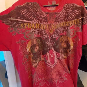 Röd t-shirt med dödskalle och vingar - Säljer en cool röd t-shirt med ett stort tryck av dödskallar, vingar och pistoler på framsidan. På baksidan finns en stor dödskalle med texten 'ARMED CUSTOM'. Perfekt för en avslappnad och edgy stil. T-shirten är kortärmad och gjord av bomull, vilket gör den bekväm att bära.